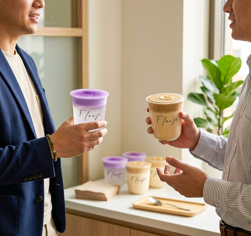 Two men in a bright office setting chatting while holding iced drinks from "Flowji" in clear plastic cups. The man on the left wears a blue blazer and holds a drink with a vibrant purple foam layer. The man on the right wears a striped button-down shirt and holds a beige, frothy drink topped with a banana slice. More Flowji drinks sit on a counter in the background.